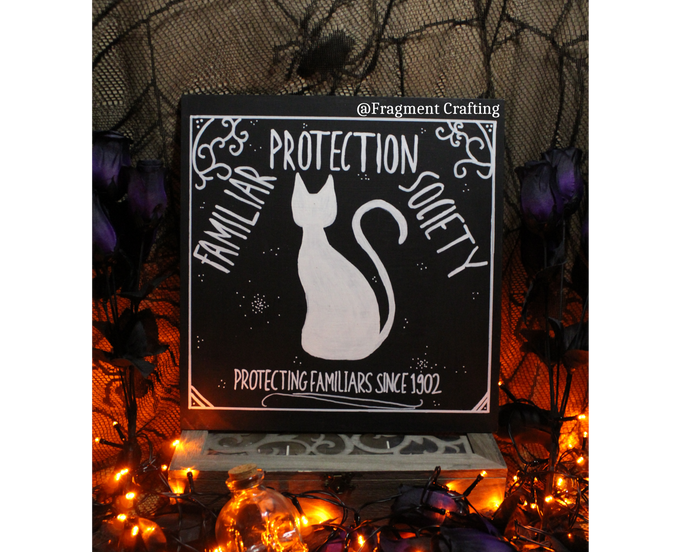 A wooden sign of a black background with a white silhouette of a cat with title of Familiar Protection Society Protecting Familiars since 1902 being showed in a Halloween setting.
