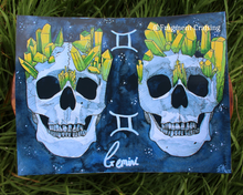 Load image into Gallery viewer, A print of a watercolour zodiac Gemini star sign of two skulls on a grey background being shown off on grass.
