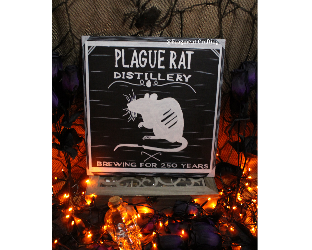 A wooden sign of a black background with a white border and rat stating Plague Rat Distillery Brewing for 250 years being showcased in a Halloween setting.