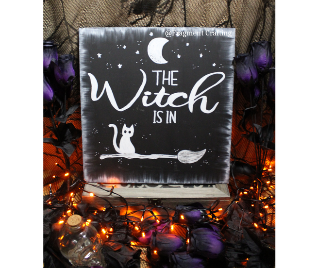 A original wooden sign of a black background with white moon and stars and cat sitting on a broom with the title The Witch Is in being showcased in a Halloween setting.
