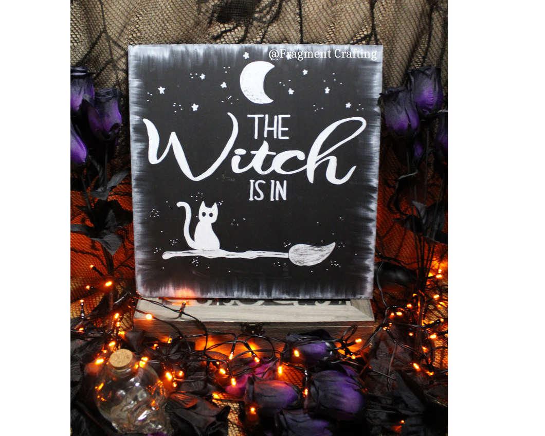 A original wooden sign of a black background with white moon and stars and cat sitting on a broom with the title The Witch Is in being showcased in a Halloween setting.